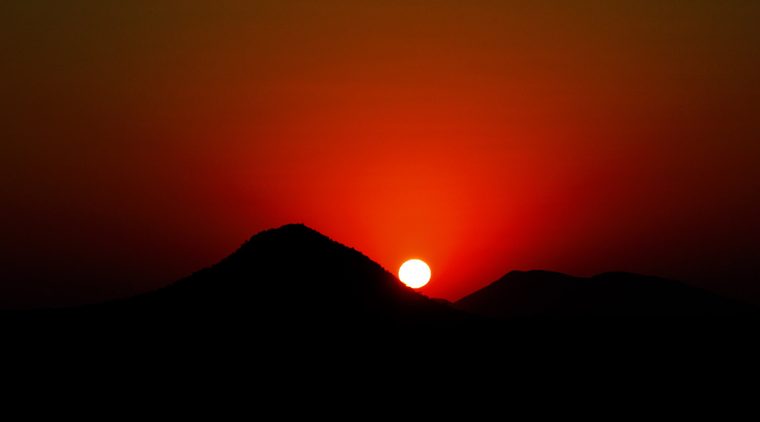 The sun sets behind a dark mountain ridge beneath a glowing red sky.