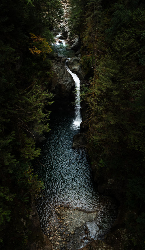 A tall waterfall drops through a narrow forest canyon into a dark pool.